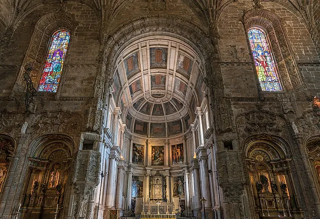 L'abside della chiesa vista dalla navata: un coro con volta a botte a cassettoni, dipinti e altare, vetrate colorate ai due lati e cappelle laterali nella luce calda