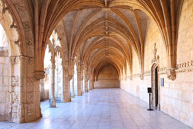Una larga galería abovedada del claustro, con techo de piedra de nervaduras, pilares tallados a la izquierda y un suelo de piedra clara que lleva hasta el fondo