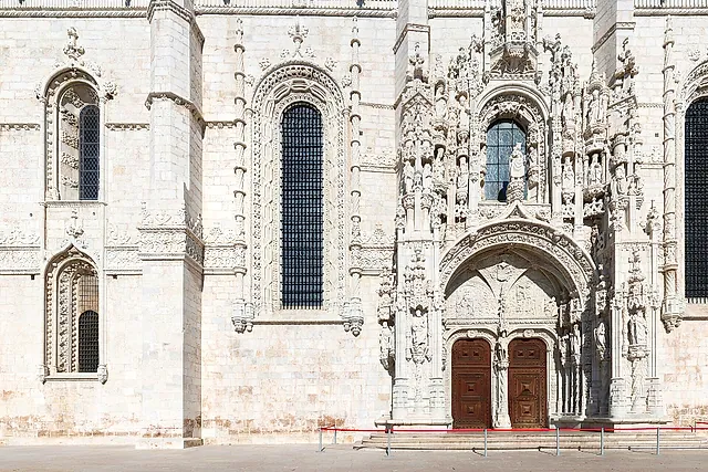 El portal sur de la iglesia del monasterio, ricamente tallado, con filas de figuras esculpidas alrededor del arco sobre dos puertas de madera oscura, junto a una alta ventana de arco en el muro de piedra clara