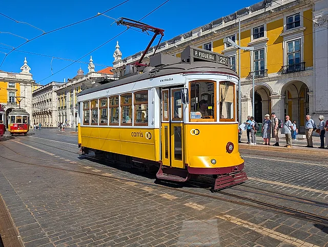 Un tranvía de época amarillo y blanco de Lisboa en la plaza empedrada de la Praça do Comércio, con los edificios amarillos con soportales detrás y algunas personas esperando en la parada, bajo un cielo azul despejado