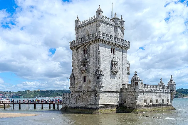 La Torre de Belém vista desde el lado del río bajo un cielo de nubes blancas, con sus torretas decoradas y un paseo con visitantes a la izquierda