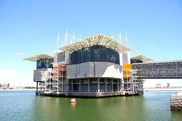 El edificio del Oceanario de Lisboa que se alza sobre el agua del dique, con cubiertas de cristal, mástiles blancos y cables sobre muros de hormigón gris, una pasarela a la derecha y una boya redonda de mascota en primer plano, bajo un cielo azul