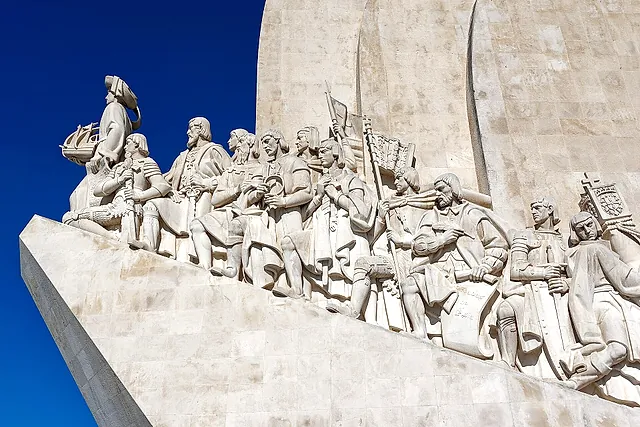 La prua scolpita del Monumento alle Scoperte, con una fila di figure di pietra guidata da una che tiene una piccola nave, contro un cielo azzurro intenso