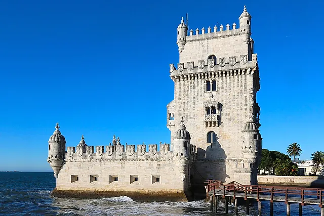 La Torre di Belém, la torre di pietra chiara con torrette e un basso bastione, sul bordo dell'acqua con una passerella di legno sotto un cielo azzurro
